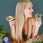 A smiling woman holding avocado and basil, surrounded by fresh vegetables and fruits for healthy eating.